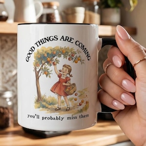 May include: White ceramic mug with a black handle and rim. The mug features a vintage illustration of a girl picking apples from a tree, with the text "Good things are coming, you'll probably miss them."
