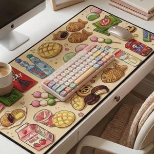 May include: A beige desk mat featuring a colorful illustration of Japanese snacks and drinks, including taiyaki, dango, and strawberry milk. A pastel keyboard and computer mouse sit on the mat. A cup of coffee and a framed quote are also visible.