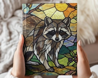 May include: A journal with a stained-glass style raccoon design. The cover features a detailed illustration of a raccoon with brown, gray, and white fur, set against a backdrop of yellow, green, and blue. The journal is held in a person's hands.