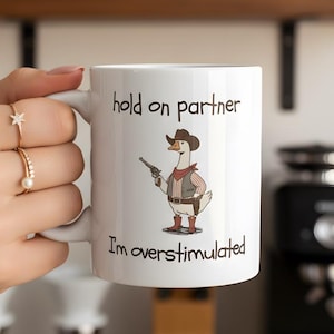 May include: White ceramic mug with the text "hold on partner" and "I'm overstimulated". The mug features a cartoon goose dressed as a cowboy holding a gun. The mug is held by a person.