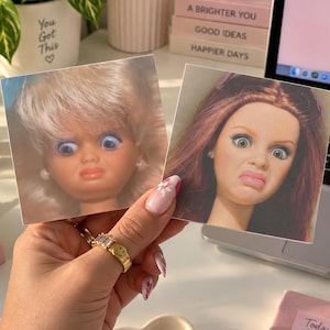 May include: Two square stickers featuring doll faces with expressive, wide-eyed, and pouty facial expressions. One sticker displays a blonde doll with blue eyes, while the other shows a brunette doll with brown eyes, both captured in a humorous, exaggerated style.