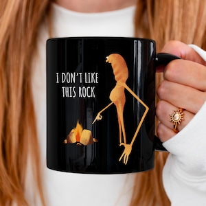 May include: Black ceramic mug with the text "I DON'T LIKE THIS ROCK" and a cartoon illustration of a figure pointing at a campfire. The figure is a light brown color.