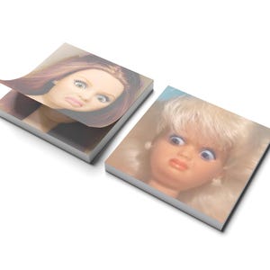 May include: Two square notepads with images of doll faces. One notepad has a page lifted, revealing a woman's face with red hair and a displeased expression. The other notepad shows a doll's face with blonde hair and a similar expression.
