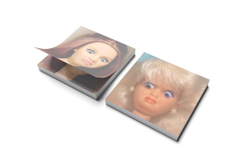 May include: Two square notepads with images of doll faces. One notepad has a page lifted, revealing a woman's face with red hair and a displeased expression. The other notepad shows a doll's face with blonde hair and a similar expression.