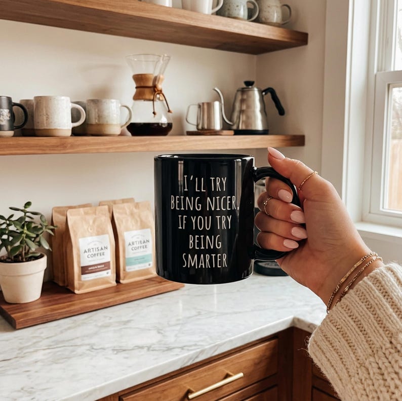 May include: A black coffee mug with the text "I'll try being nicer if you try being smarter" in white. The mug is held in a hand with several rings. Coffee beans and other coffee-making items are in the background.