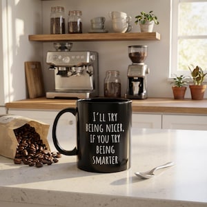 May include: A black coffee mug with the text "I'll try being nicer, if you try being smarter." The mug is on a white countertop next to a bag of coffee beans and a spoon. A coffee machine and grinder are in the background.
