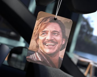 May include: Rectangular air freshener featuring a smiling portrait. The image shows a person with light brown hair and a striped shirt. The air freshener is hanging in a car, suspended by a white string.