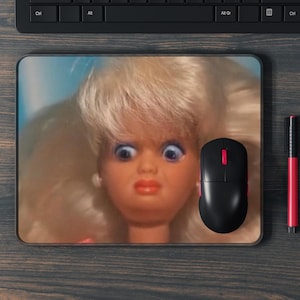 May include: A rectangular mousepad featuring a doll's face with wide eyes and a surprised expression. A black computer mouse with a red accent and a red and black pen are next to the mousepad. The background is a dark wood grain.