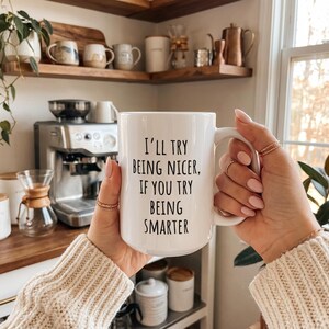 May include: A white ceramic mug with the text "I'll try being nicer, if you try being smarter" in black. The mug is held by a person with rings on their fingers. The background includes a coffee machine and other kitchen items.