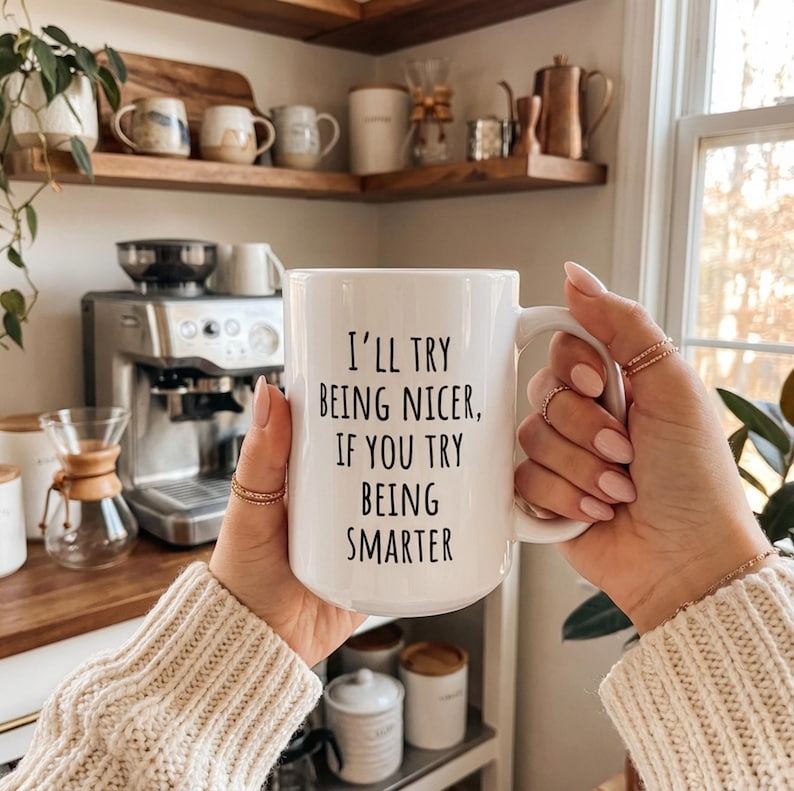 May include: A white ceramic mug with the text "I'll try being nicer, if you try being smarter" in black. The mug is held by a person with rings on their fingers. The background includes a coffee machine and other kitchen items.