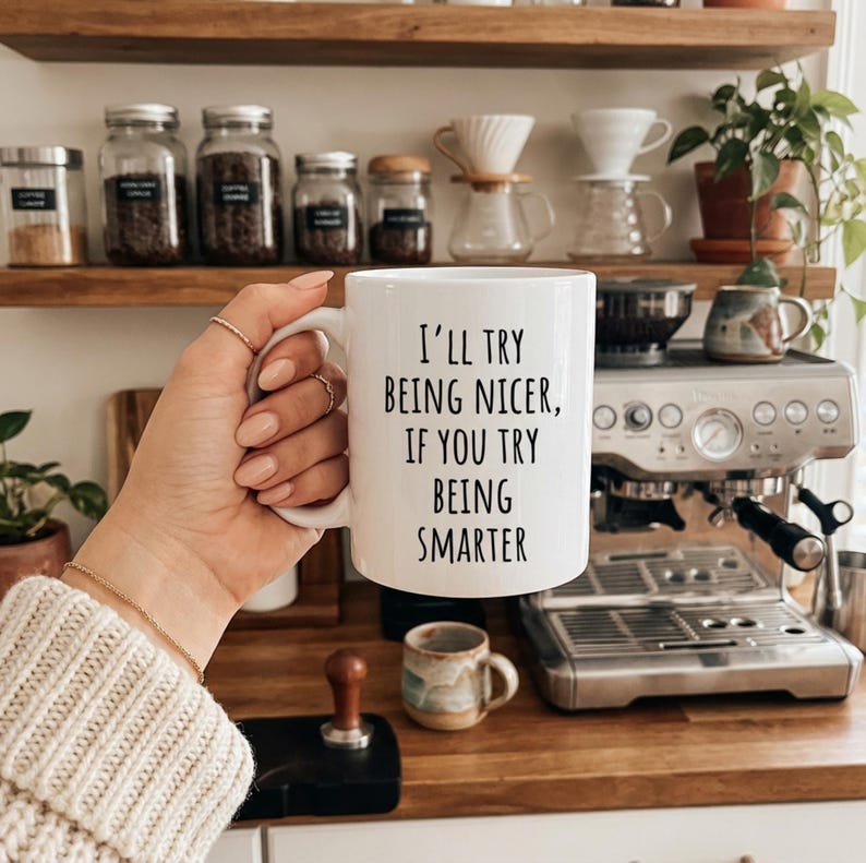 May include: A white ceramic mug with the text "I'll try being nicer, if you try being smarter" in black. The mug is held in a hand with rings and a bracelet, with a coffee station in the background.