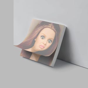 May include: A stack of white sticky notes with a printed image of a doll's face with a surprised expression. The doll has long brown hair, large eyes, and a slightly pursed mouth.