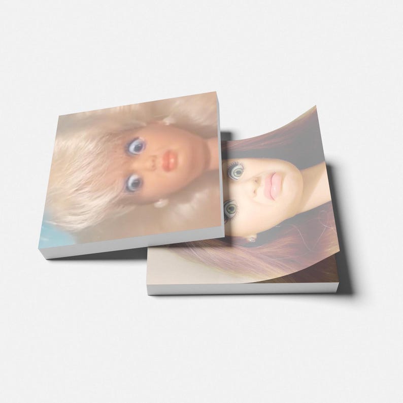 May include: Two stacked square notepads with images of doll faces. The top notepad features a doll with blonde hair and blue eyes. The bottom notepad shows a doll with brown hair and large eyes. The notepads are on a white surface.