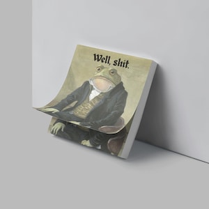 May include: A notepad with a humorous illustration of a frog in formal attire, with the text "Well, shit." The frog is depicted in a seated position, wearing a suit and waistcoat. The notepad has a white edge.