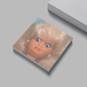 May include: A vintage doll's head with blonde hair and blue eyes, mounted on a square, white surface. The doll has a surprised expression with a slightly open mouth and red lips. The background is a light gray.