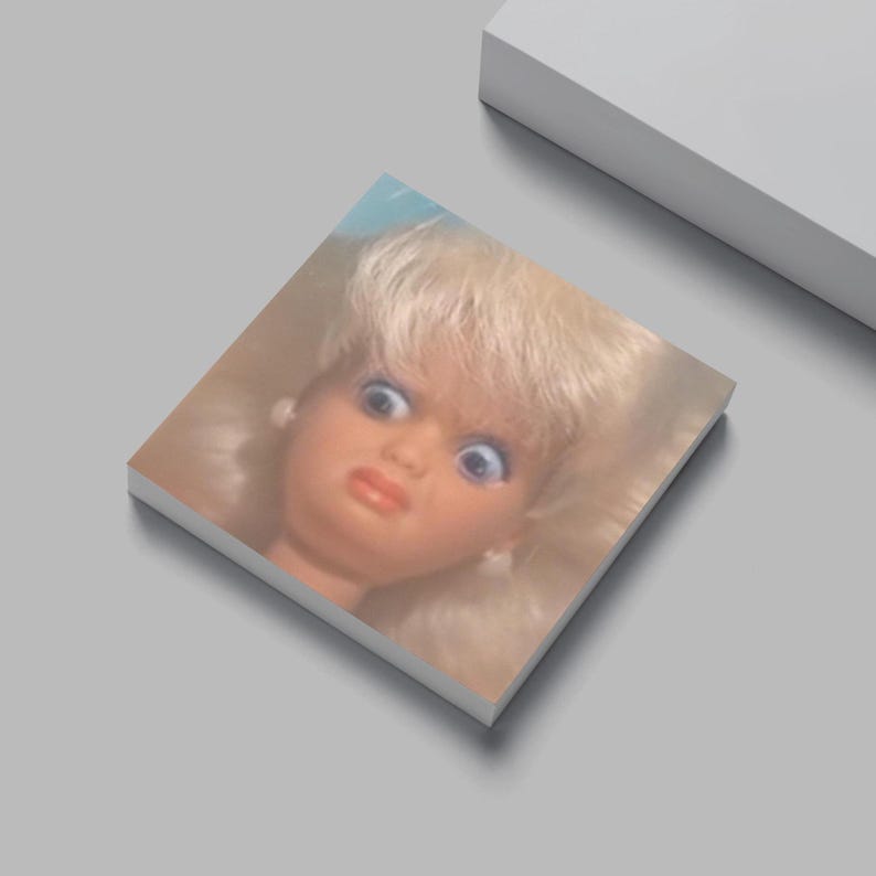 May include: A vintage doll's head with blonde hair and blue eyes, mounted on a square, white surface. The doll has a surprised expression with a slightly open mouth and red lips. The background is a light gray.