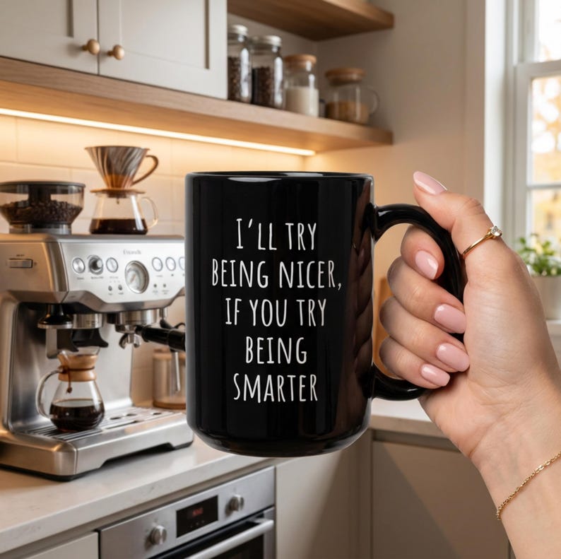 May include: Black ceramic coffee mug with the text "I'll try being nicer, if you try being smarter" in white. The mug is held by a person. A coffee machine and coffee-making equipment are in the background.