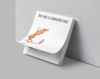 May include: A white notepad with the text "THIS PLACE IS A DANGEROUS PLACE" and a cartoon illustration of a shrimp-like creature. The notepad is propped up against a gray wall.