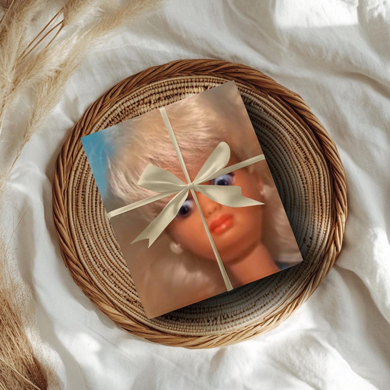 May include: A wrapped gift featuring a doll's face, tied with a cream-colored ribbon, rests on a woven basket. The doll has blonde hair, blue eyes, and red lips. The basket sits on a white fabric surface.