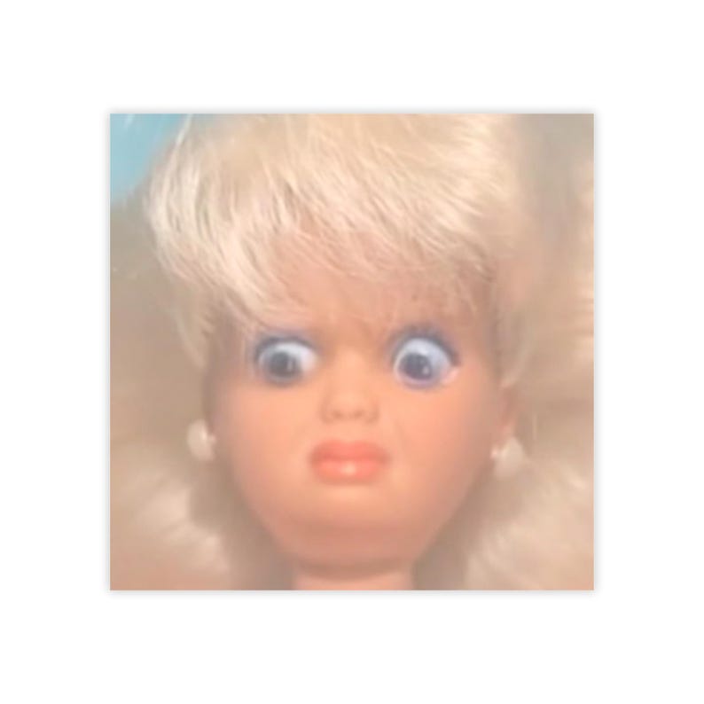 May include: A close-up of a doll's face with wide, blue eyes and a surprised expression. The doll has blonde hair and a pale complexion. The doll's lips are a bright red color.