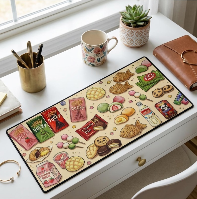 Puede incluir: Alfombrilla de escritorio rectangular con un fondo beige decorado con ilustraciones coloridas de snacks japoneses. El dise&ntilde;o incluye cajas de Pocky, barras Kit Kat, pasteles con forma de pez y mochi. La alfombrilla tiene un borde negro.