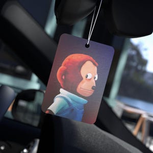 May include: Rectangular car air freshener featuring a cartoon monkey with red fur and a blue denim outfit. The air freshener is hanging from a string inside a car, with a blurred background of the car's interior.