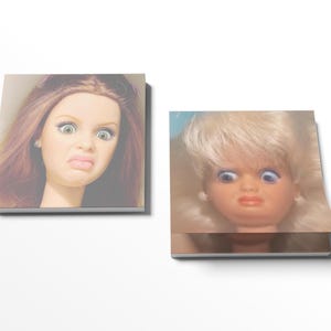 May include: Two square notepads featuring doll faces with exaggerated expressions. One doll has auburn hair and green eyes, the other has blonde hair and blue eyes. Both dolls have a surprised or disgusted expression.