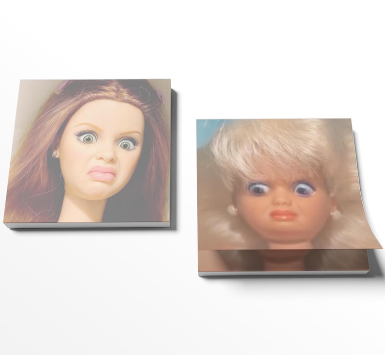 May include: Two square notepads featuring doll faces with exaggerated expressions. One doll has auburn hair and green eyes, the other has blonde hair and blue eyes. Both dolls have a surprised or disgusted expression.