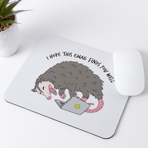 May include: White mousepad with a cartoon opossum illustration. The opossum is gray with a pink nose and tail, and is lying next to a laptop with a sad face on the screen. The text "I HOPE THIS EMAIL FINDS YOU WELL" is above the opossum.