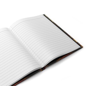 May include: An open lined journal with a decorative cover. The pages are white with gray lines. The cover has a dark, patterned design with shades of brown, black, and gold. The journal is angled, showing the interior pages and the cover's edge.