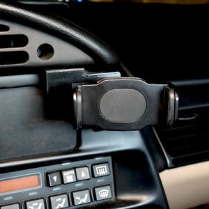 May include: A black car phone holder mounted on a dashboard. The holder is attached to a vent and has an adjustable grip to secure a phone. The dashboard is black with various buttons and vents.