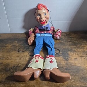 May include: A vintage cowboy marionette puppet with a painted face, red hair, and freckles. The puppet wears a plaid shirt, blue pants, and brown cowboy boots with white cuffs. The boots have a red star and the letters "HD" on them.