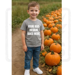 May include: A young child wearing a grey t-shirt with the text "YOUR RAD DESIGN GOES HERE" in white, blue jeans, and white trainers. The child smiles in front of a pumpkin patch with many orange pumpkins.