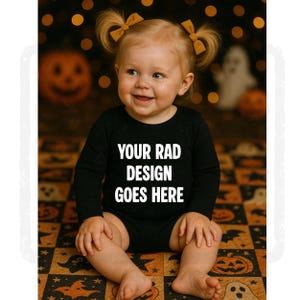 May include: A smiling baby wearing a black long-sleeved onesie with the text "YOUR RAD DESIGN GOES HERE." The baby has blonde hair styled in pigtails with yellow bows. The background features Halloween-themed decorations.