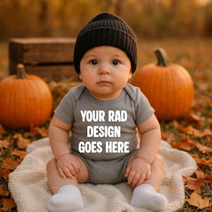 May include: A baby wearing a gray onesie and a black beanie sits on a cream-colored blanket. The onesie has the text "YOUR RAD DESIGN GOES HERE" in white. Two orange pumpkins and fall foliage are in the background.