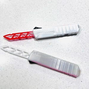 May include: Two white and red knives with a textured handle and a blade with a geometric design. The knives are lying on a light-colored surface. One knife has a red blade, and the other has a clear blade.