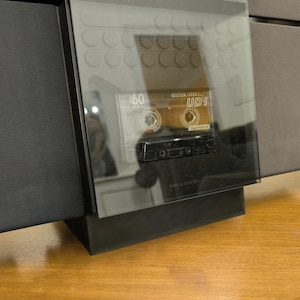 May include: A black audio cassette player with a clear cover, displaying a vintage cassette tape labelled "UDI". The player is set on a wooden surface, with a dark, rectangular base and a larger, black structure behind it.