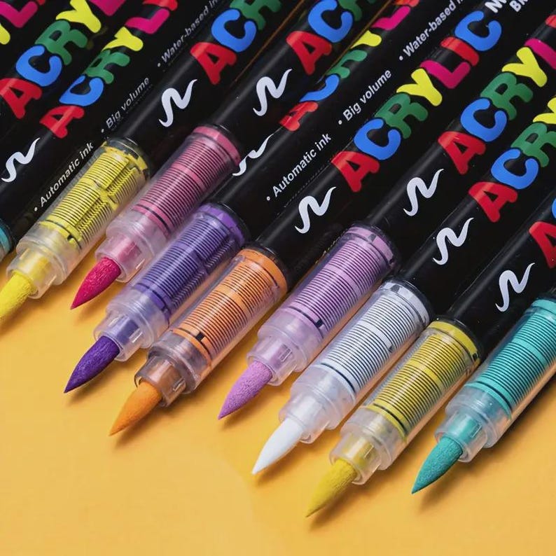 May include: A collection of colorful acrylic paint markers with black barrels. Each marker has a different colored tip, including yellow, pink, purple, orange, white, and teal. The markers are labeled with the word "ACRYLIC" in various colors.