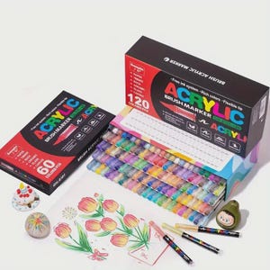 May include: An assortment of acrylic brush markers in a variety of colors. The image shows two boxes of markers, one with 60 colors and the other with 120 colors. The boxes are red and black with the word "ACRYLIC" in large letters. Several markers are lying on a white surface next to a drawing of flowers.