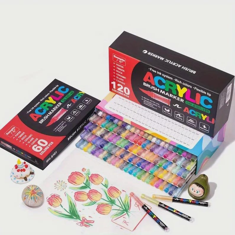 May include: An assortment of acrylic brush markers in a variety of colors. The image shows two boxes of markers, one with 60 colors and the other with 120 colors. The boxes are red and black with the word "ACRYLIC" in large letters. Several markers are lying on a white surface next to a drawing of flowers.