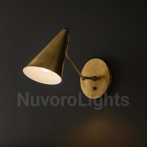 May include: A brass-toned wall sconce with a conical shade and a circular backplate. The sconce has a curved arm and a bright light bulb inside the shade. The text "NuvoroLights" is visible at the bottom.