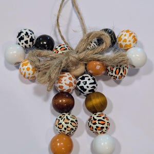 May include: A decorative beaded ornament featuring a variety of wooden beads in black, white, brown, and orange. Some beads have animal print patterns. The beads are strung together with a jute twine bow and hanging loop.