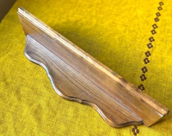 May include: A wooden shelf with a decorative, scalloped edge. The shelf is made of light brown wood and has a natural wood grain. The shelf is angled and appears to be mounted on a wall. The background is a yellow fabric.