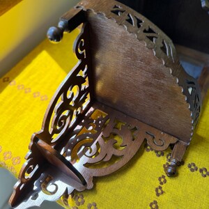 May include: A dark brown wooden corner shelf featuring intricate scrollwork cutouts along the side panels. The shelf has a flat top surface and is supported by small, rounded wooden feet at the base, showcasing a detailed fretwork design.