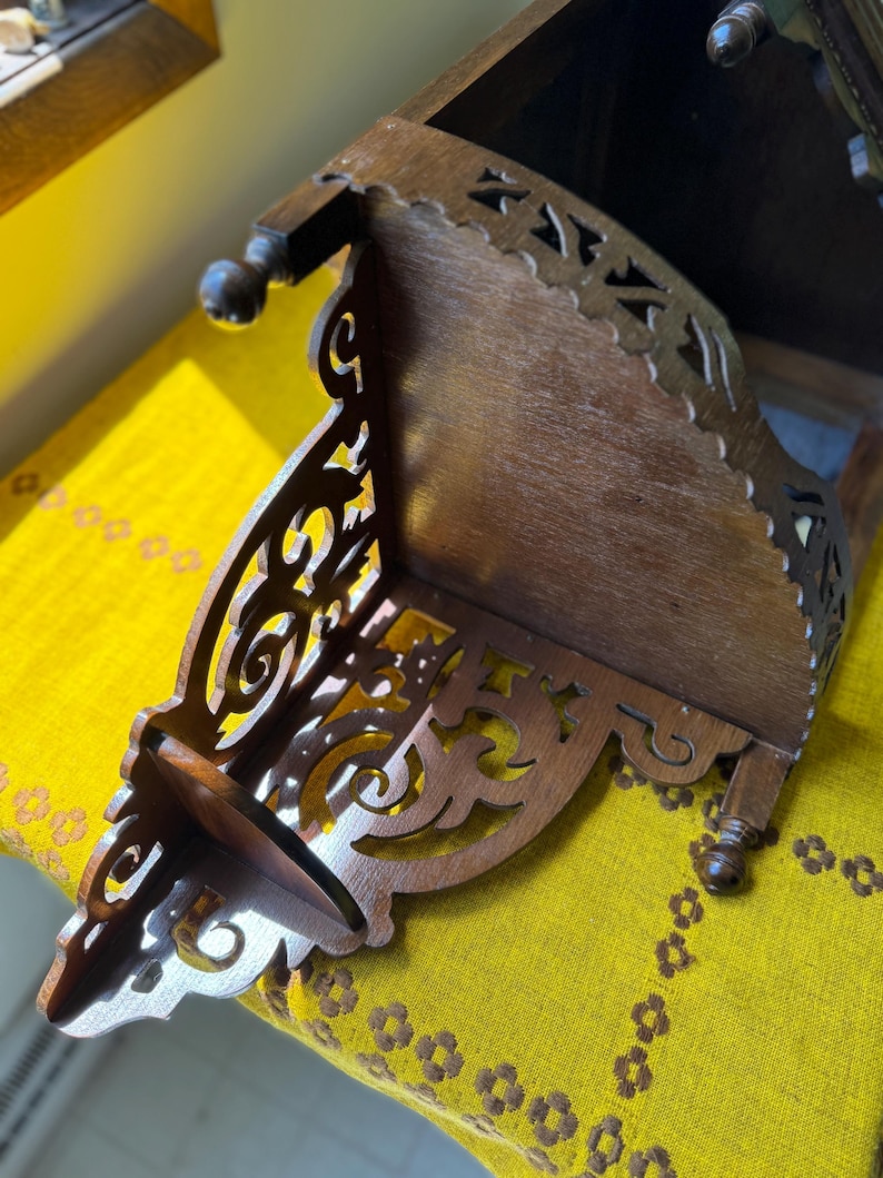 May include: A dark brown wooden corner shelf featuring intricate scrollwork cutouts along the side panels. The shelf has a flat top surface and is supported by small, rounded wooden feet at the base, showcasing a detailed fretwork design.