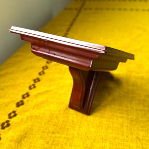 Puede incluir: Una pequeña estantería de madera teñida oscura con un soporte decorativo. La estantería es de un rico color marrón rojizo y parece estar hecha de madera maciza. La estantería está diseñada para montarse en la pared y es adecuada para exhibir objetos pequeños.
