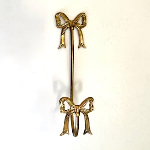 May include: A gold-colored metal bow-shaped wall hook. The hook has two decorative bows, one at the top and one at the bottom, connected by a vertical rod. The bows have detailed, textured designs.