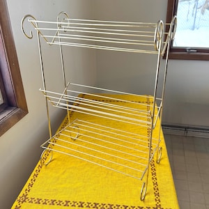 May include: A three-tiered, gold-colored metal shelf. The shelf has a rectangular shape with horizontal bars for each level. The frame has decorative scrollwork on the sides and feet. The shelf is displayed on a yellow tablecloth.