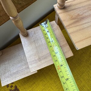 May include: A wooden, tiered display stand with a tape measure showing measurements in inches. The stand has a light wood finish and a turned leg design. The tape measure shows a measurement of approximately 4 inches.