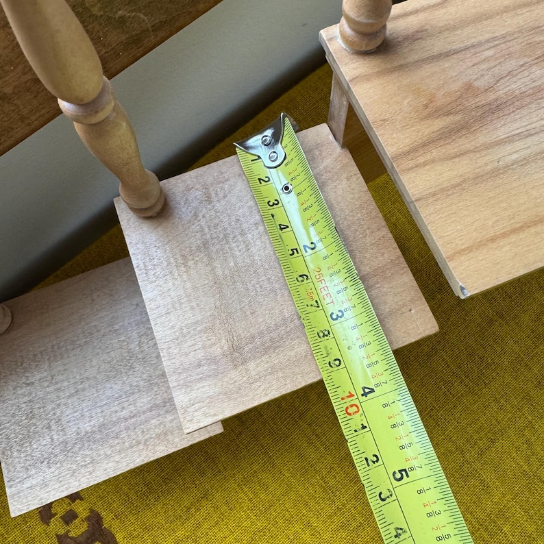 May include: A wooden, tiered display stand with a tape measure showing measurements in inches. The stand has a light wood finish and a turned leg design. The tape measure shows a measurement of approximately 4 inches.